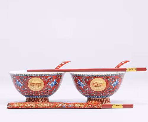 Two red ceramic bowls with gold double-happiness medallions and blue floral motifs, each resting on a red brocade chopstick pouch with red chopsticks and spoons