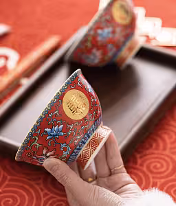 Hand holding a red ceramic bowl close-up, showing gold double-happiness medallion and intricate blue lotus scrollwork on the exterior