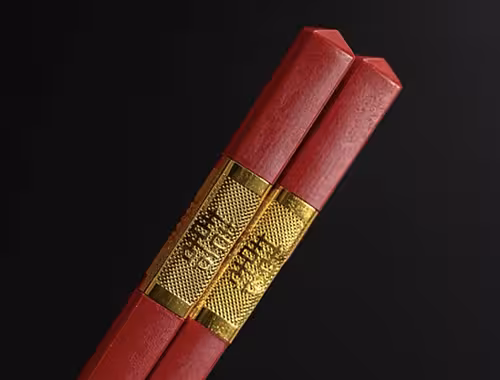 Close-up of two red wooden chopsticks with an engraved gold band featuring Chinese characters, set against a dark background