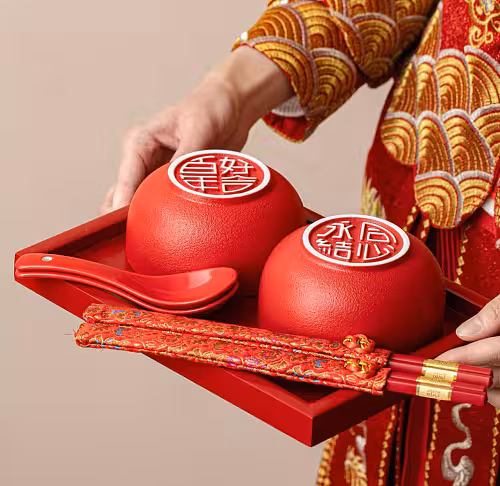 Person in red gold embroidered qipao holding a red tray with two matte red bowls, red spoons, and brocade-wrapped chopsticks