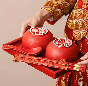 Person in red gold embroidered qipao holding a red tray with two matte red bowls, red spoons, and brocade-wrapped chopsticks