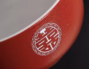Macro shot of a red ceramic bowl rim showing a white embossed double happiness medallion against the textured matte red surface