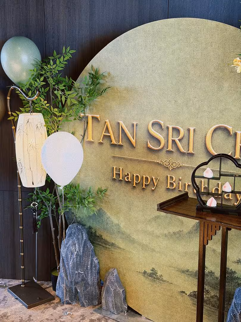 Round sage green mountain-landscape birthday backdrop flanked by a bamboo zen floor lamp, artificial nandina tree, decorative rocks, and sage balloons