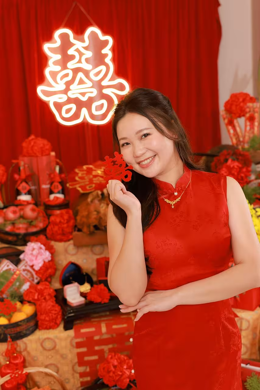 A bold red-and-gold traditional Chinese wedding setting centred around the glowing 囍 Double Happiness LED Neon Light Signage, hung against rich red draping. The foreground is layered with red carnation blooms, fresh oranges, pomegranates, gold-patterned gift boxes, and festive red decorative accents, creating a warm and auspicious atmosphere.