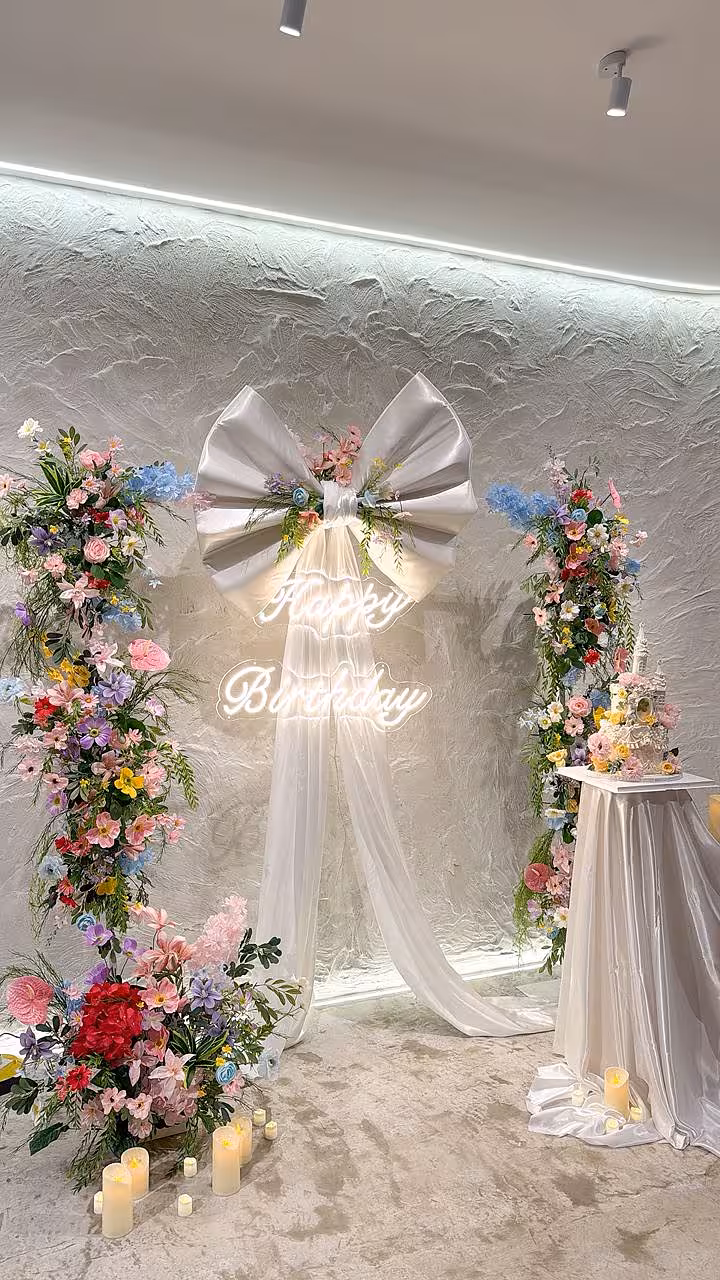A dreamy backdrop features two lush floral columns in pink, blue, purple, and red blooms flanking a glowing Happy Birthday LED Neon Light Signage, topped with an oversized silver satin bow and cascading white drape. Pillar candles on the floor and a flower-adorned cake on a draped pedestal table complete the soft, garden-party mood.