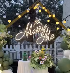 Marry Me LED Neon Light Signage glowing warm white at an outdoor dusk proposal setup with string lights, green balloon arch, and floral table centrepiece