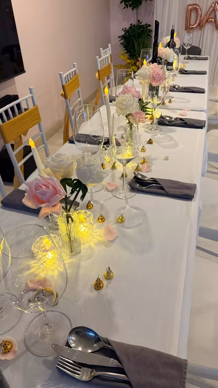 A white linen long table is dressed with clusters of pink and white artificial curl-edge roses in assorted mini clear glass vases, warm-glowing LED taper candles in glass pedestal holders, and sparkling crystal-effect tealight cups that cast a soft golden glow across the tablescape. Grey folded napkins, polished silverware, and scattered rose petals complete the romantic, intimate setting.