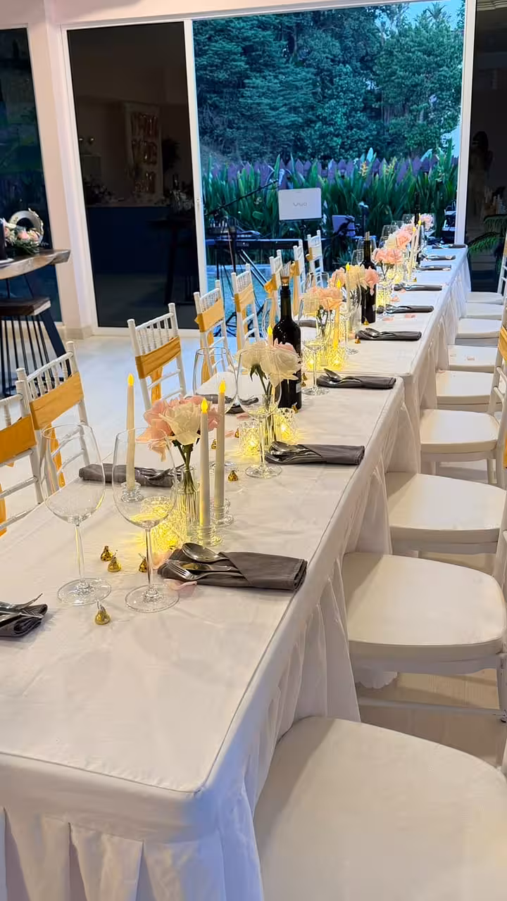 A dreamy long-table setting dressed in white linen, featuring clusters of blush pink and white artificial curl-edge roses in mini clear glass vases, slender LED taper candles on glass European pedestal holders, and softly glowing crystal-effect tealight cups that cast a warm amber shimmer across the tablescape.