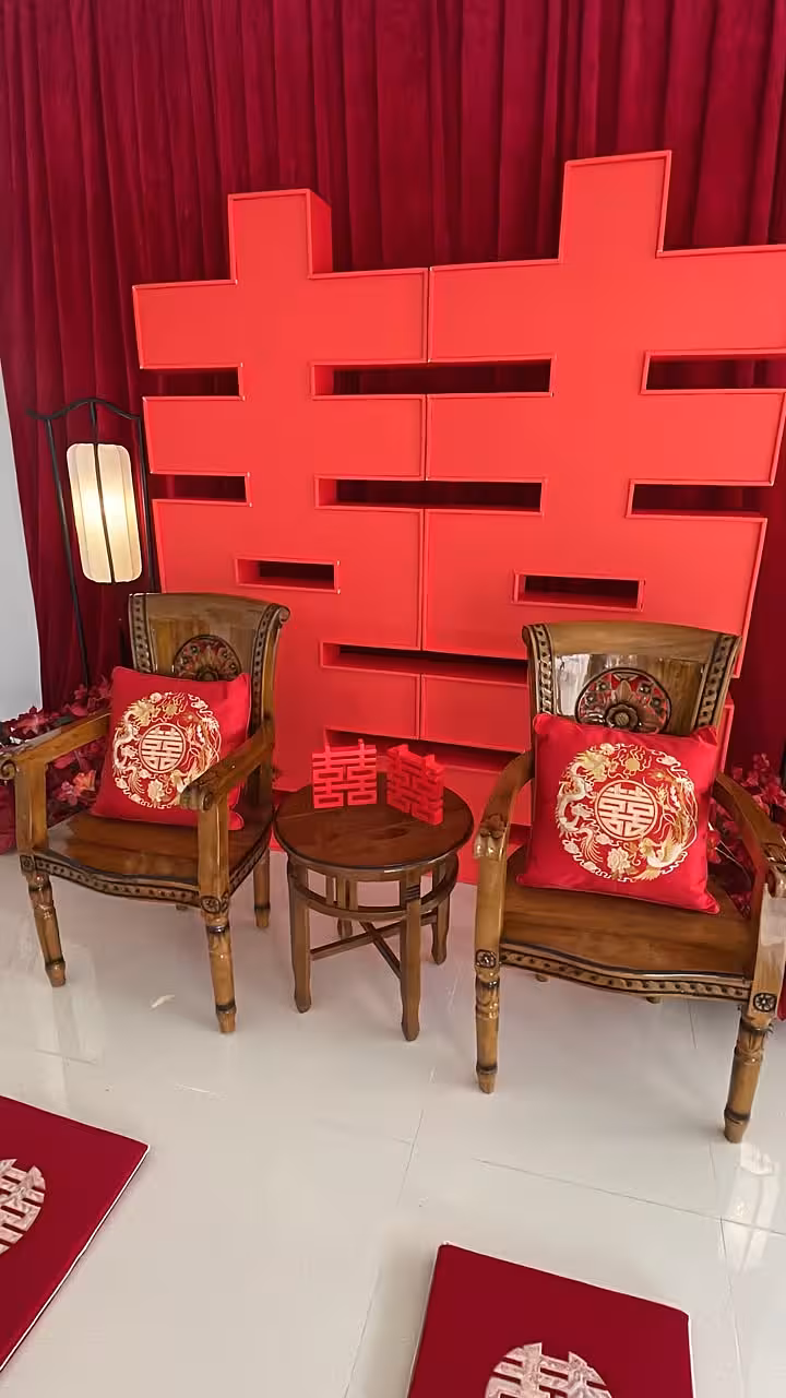 A rich crimson palette anchors this traditional Chinese wedding vignette, featuring a bold 2-metre 囍 Double Happiness backdrop set against a wine red velvet curtain, framed by a retro arch lantern floor lamp on one side and red artificial floor flowers on the other. Two carved wooden chairs dressed with gold-embroidered dragon and phoenix cushion covers flank a small round table topped with a decorative 囍 motif prop.