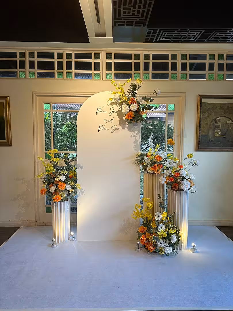 A crisp white arch backdrop with personalised script signage is framed by lush floral clusters in warm orange, yellow, and white tones. Three Structure Origami Cylindrical Pleated Flower Desserts Stands in staggered heights anchor the arrangement, lit softly from below against a white carpet floor.
