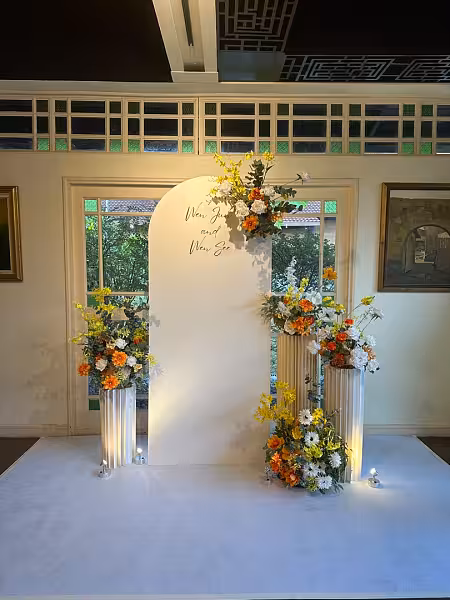 White arch signage backdrop flanked by three white origami cylindrical pleated stands topped with orange, yellow, and white floral arrangements