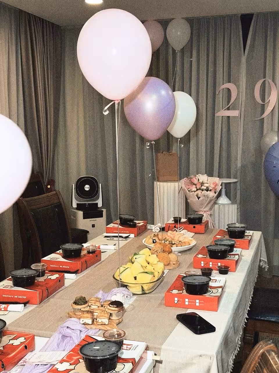 Soft pink, lilac, and white helium balloons float above a long table dressed with a neutral runner, while origami cylindrical pleated dessert stands anchor the spread in the background. Large blush number signage adorns the grey curtain backdrop alongside a wrapped floral bouquet, creating a dreamy, muted-toned celebration setup.