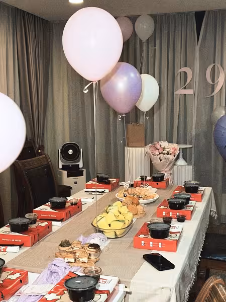 Pastel pink, lilac, and white balloon clusters above a long dining table styled with origami cylindrical pleated dessert stands, number signage, and a floral bouquet against grey curtains
