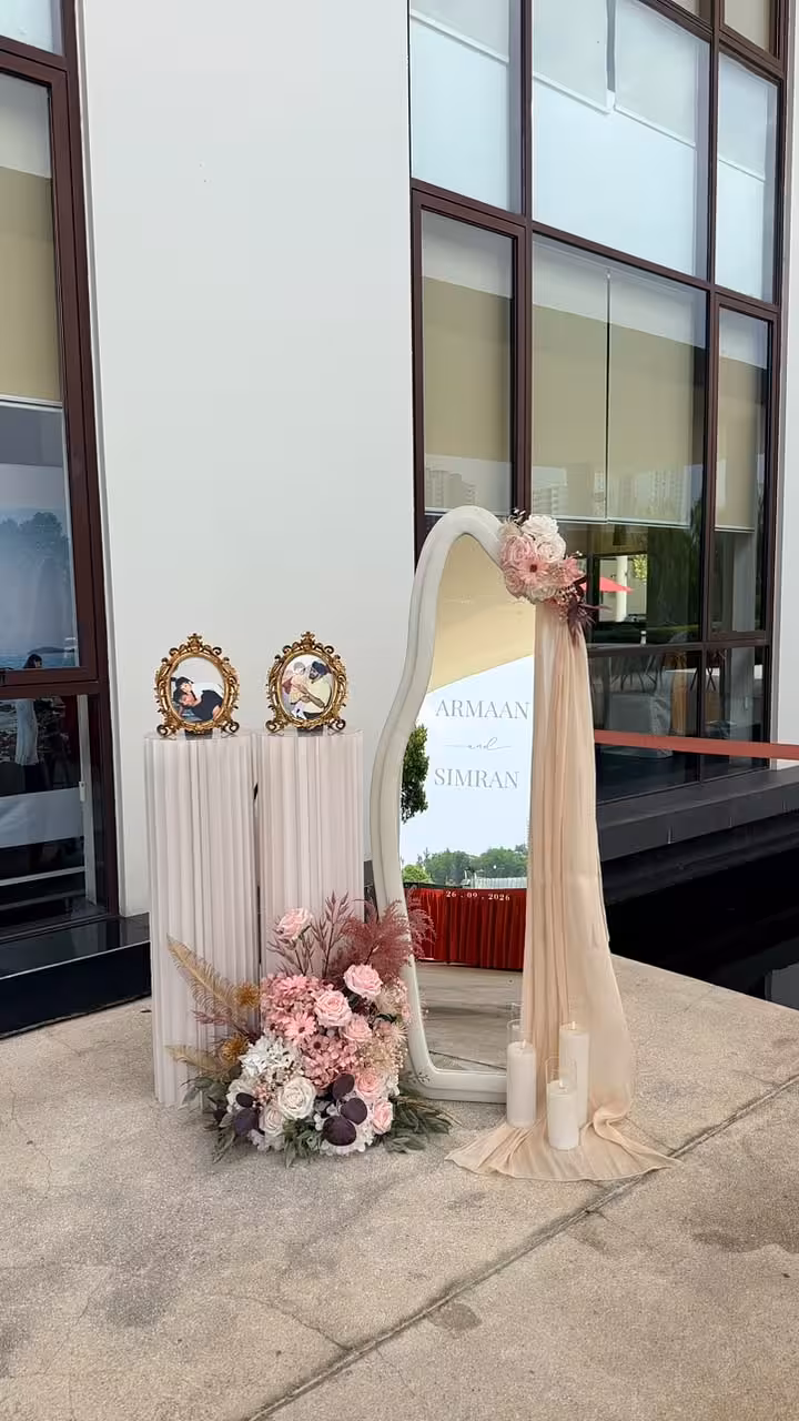 Two Structure Origami Cylindrical Pleated Flower Dessert Stands in soft white are topped with ornate gold-framed pieces, flanking a wavy full-length mirror adorned with blush and burgundy blooms and a flowing champagne chiffon drape. A lush ground floral arrangement of pink roses, dried pampas grass, and deep plum flowers anchors the vignette alongside pillar candles.