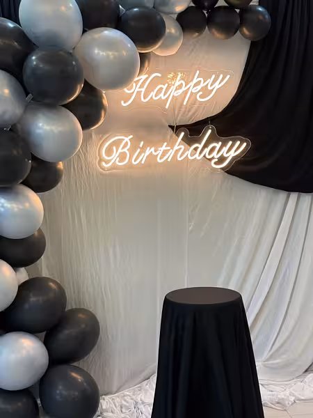 Happy Birthday LED neon light sign in warm white cursive against a white drape backdrop with black and grey balloon garland