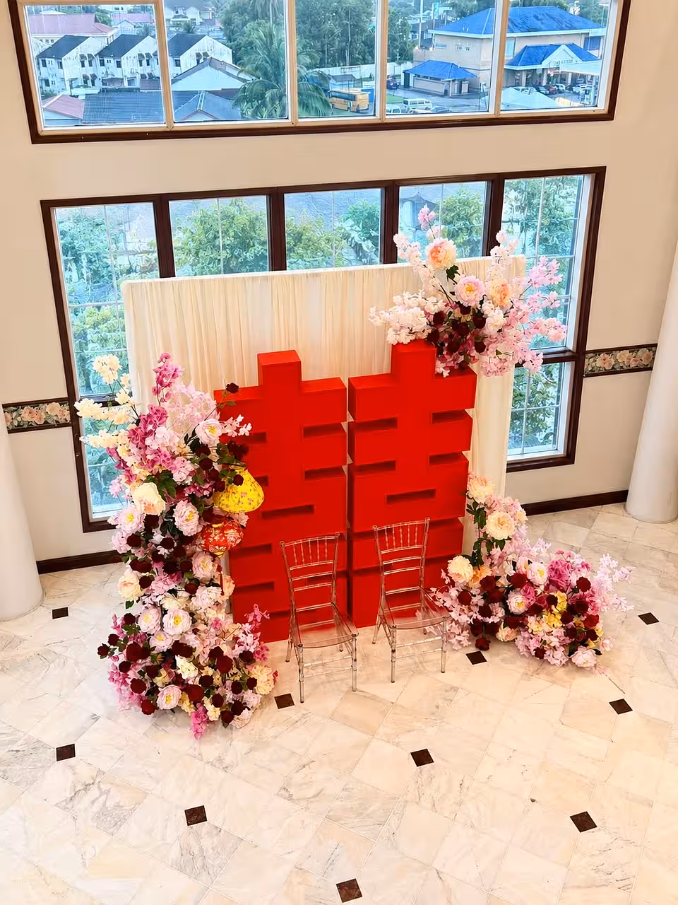 A bold 2-meter red 囍 Double Happiness character takes centre stage against a soft cream drape backdrop, flanked by lush cascading floral arrangements in shades of blush pink, white, and deep burgundy. A traditional red lantern accent and clear Chiavari chairs complete the elegant, festive vignette.