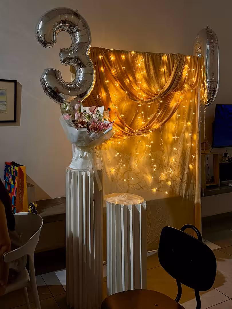 A luxuriously draped champagne fabric backdrop glows warmly against cascading LED fairy lights, creating a golden, intimate ambience. In front, two white origami-pleated cylindrical dessert stands of varying heights are topped with a floral bouquet and a decorative bust, while oversized silver number balloons float above the scene.