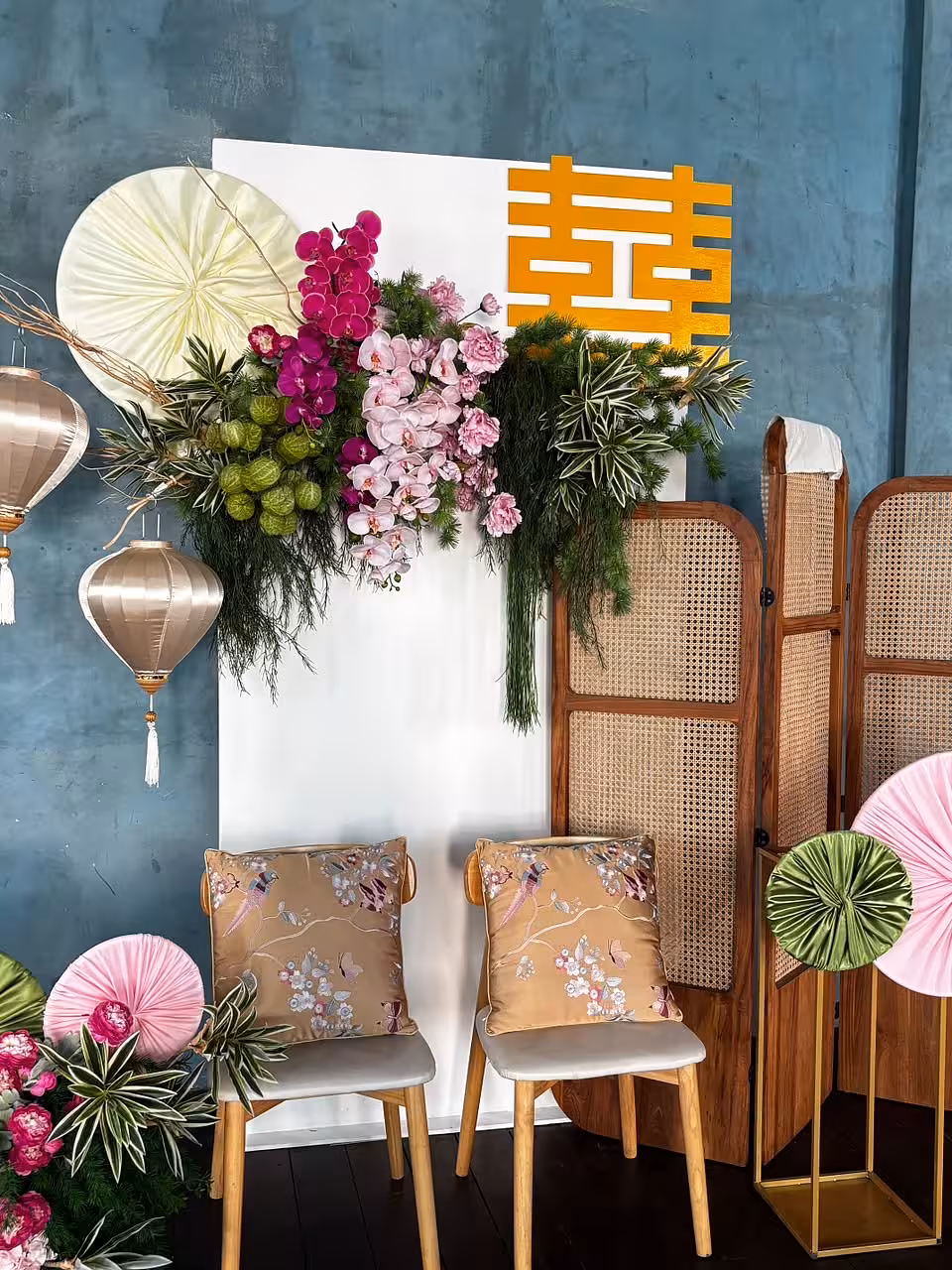A modern Oriental-inspired vignette features a white panel backdrop adorned with lush magenta and blush orchids, greenery, and a bold gold double happiness character. Silk lanterns, rattan screens, paper fan discs, and chairs dressed with gold Oriental Embroidered Floral Bird Butterfly Designed Decorative Satin Cushions complete the warm, eclectic scene.