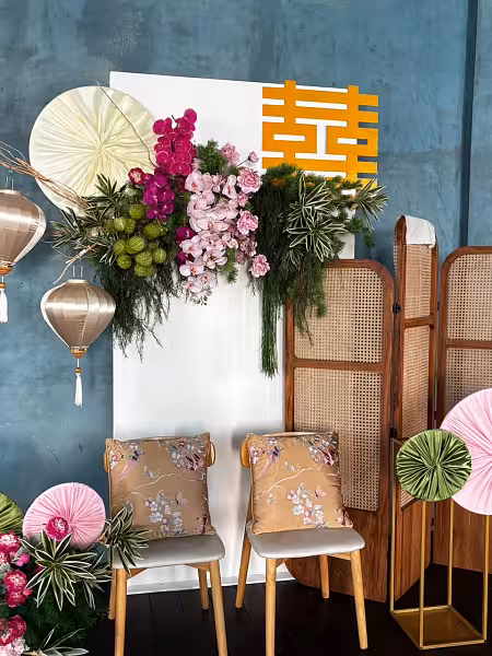 White panel backdrop with pink and magenta orchid floral arrangement, gold double happiness sign, rattan room divider, silk lanterns, and embroidered satin cushions on wooden chairs