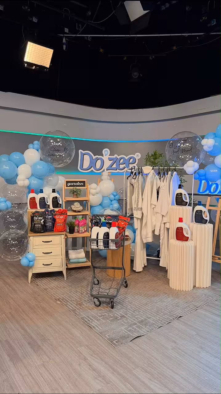 A lively branded studio display styled with a sweeping blue and white organic balloon garland, large clear orb balloons with printed text, and a trio of white origami cylindrical pleated dessert stands in varying heights. A tall wooden easel stand anchors one side of the clean, airy scene.