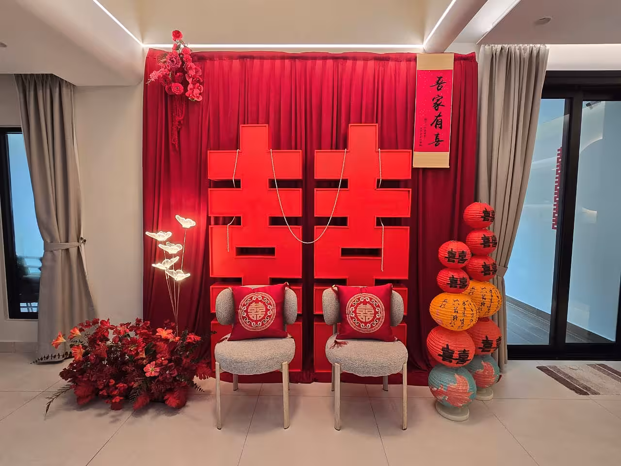 Red 囍 Double Happiness backdrop with stacked paper lanterns, butterfly LED floor lights, red artificial flowers, and embroidered cushion chairs
