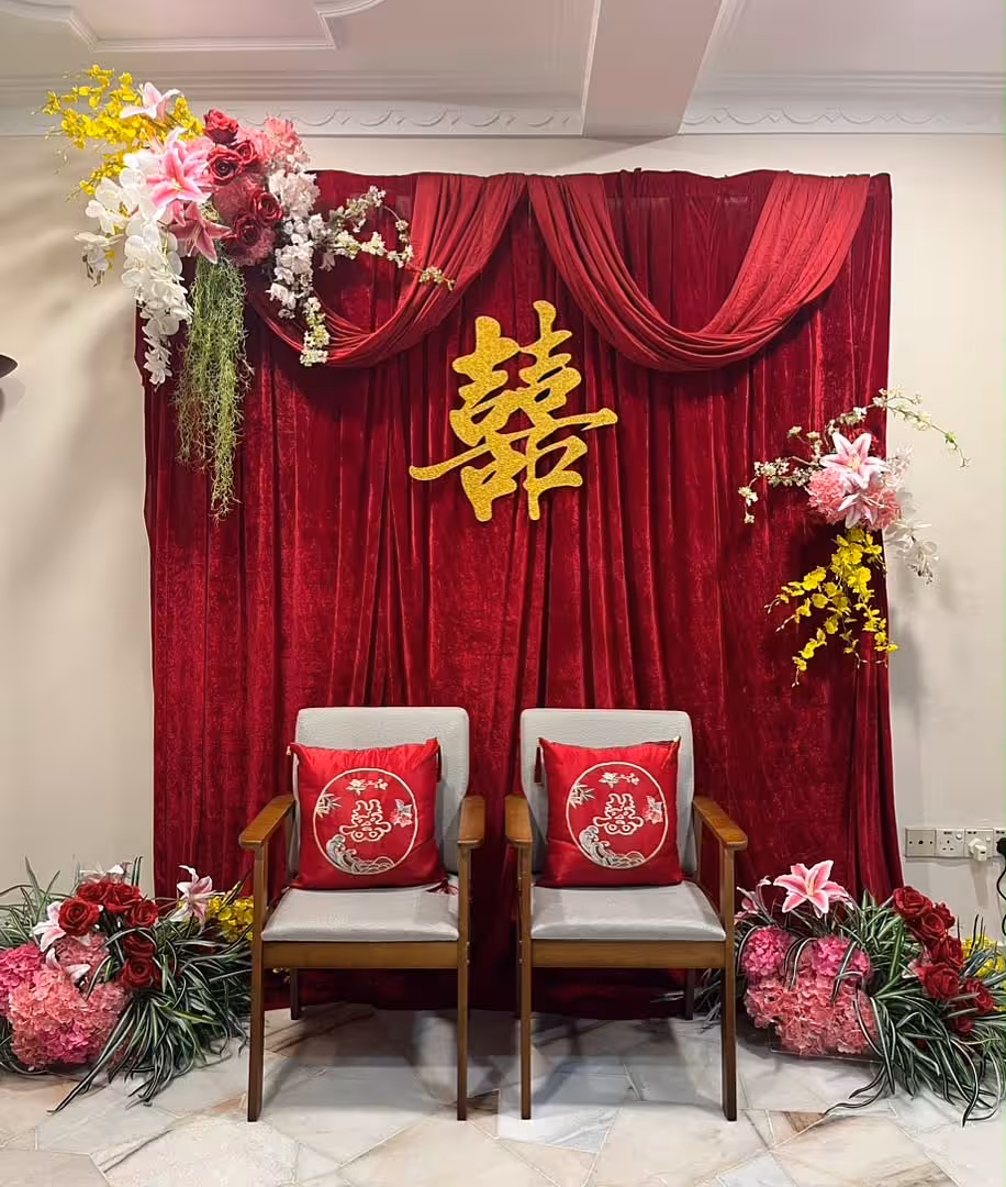 Rich red velvet draped backdrop with large gold double-happiness ornament, flanked by colourful artificial flowers and two walnut wooden chairs with red embroidered dragon and phoenix cushions