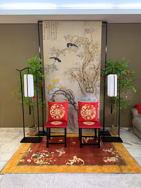 Black wooden chair set with dragon and phoenix embroidered red cushions on a vibrant oriental carpet, framed by a Chinese ink painting backdrop, arch lantern floor lamps, and artificial nandina trees