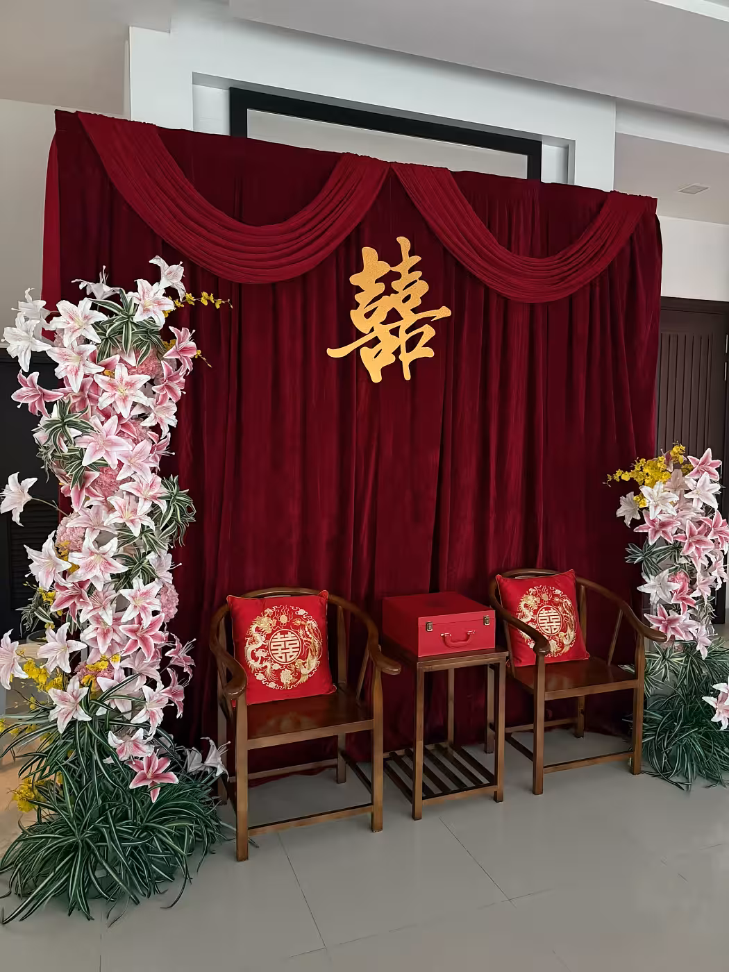 Wine red velvet draped backdrop with gold Xi ornament, flanked by pink lily flower arches and walnut chairs with dragon phoenix cushions