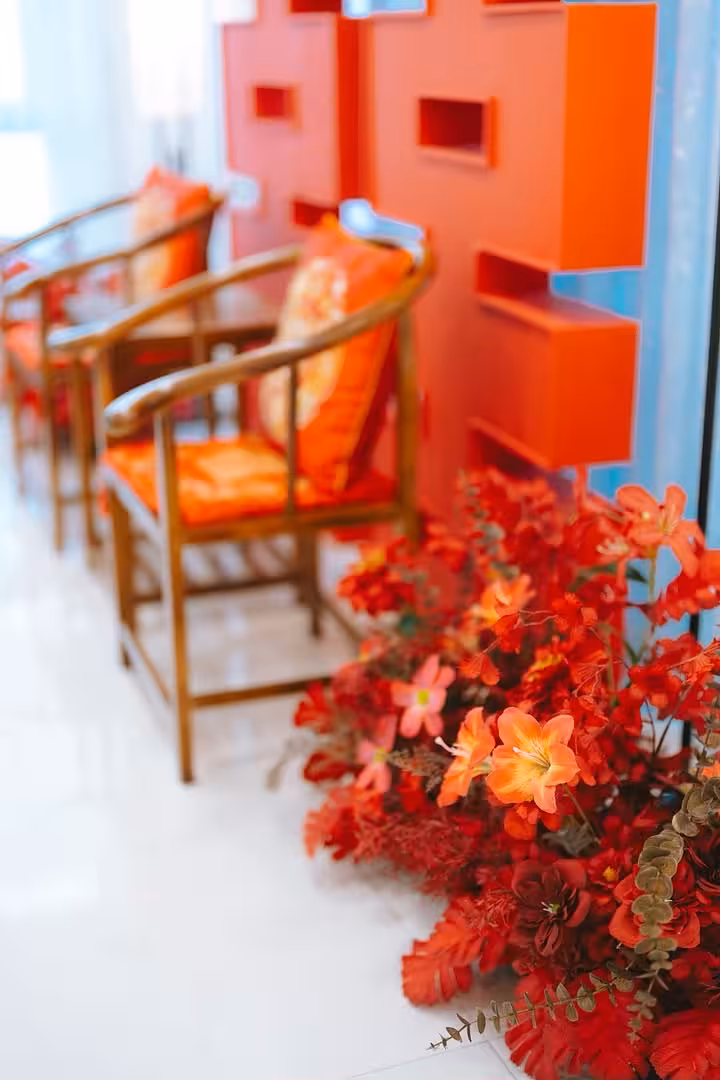 Bloom Red Artificial Floor Flowers arranged beside a row of wooden armchairs with orange cushions and a bold red geometric backdrop