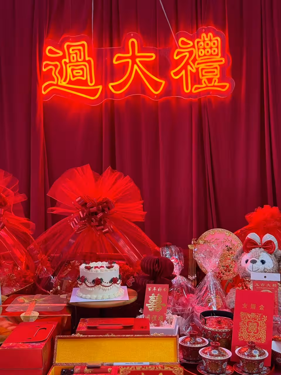 A bold wine red velvet curtain backdrop anchors the setup, with a glowing 过大礼 neon light sign in gold and red suspended above. The table below is richly layered with traditional betrothal gifts, red ribbon fan decorations, auspicious signage, and ceremonial items, creating a vibrant and festive GuoDaLi atmosphere.