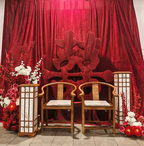 Wine red velvet decorative backdrop curtain with a large fabric double happiness symbol, two wooden Ming-style chairs, tall shoji lanterns, and red, pink, and white floral arrangements