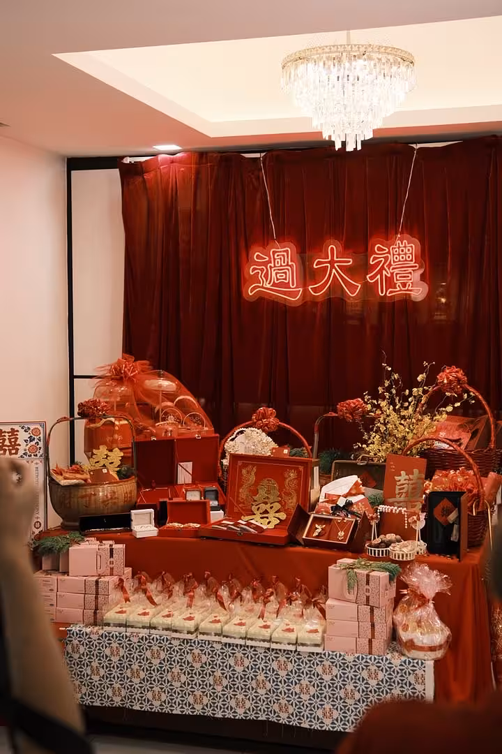 A rich wine red velvet curtain backdrop, hung on a black metal arch stand, frames a glowing 过大礼 neon light signage in warm white and red. Below, a satin-draped betrothal display table is layered with traditional red gift boxes, lacquerware, gold floral arrangements, and double-happiness motif accents, creating an opulent and festive GuoDaLi atmosphere.